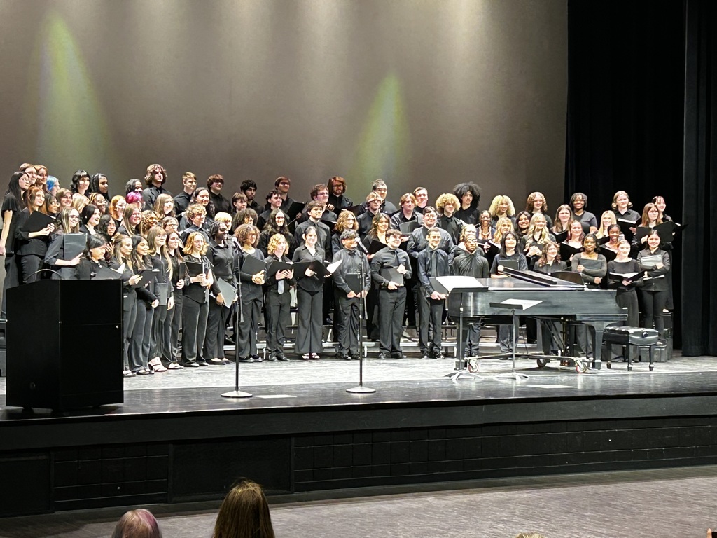 A large high school honors chorus stands on risers on stage at the Citizens Center wearing formal black concert attire. Students hold black music folders and smile toward the audience. A grand piano sits at the front of the stage, and stage lighting creates a soft green glow on the backdrop.