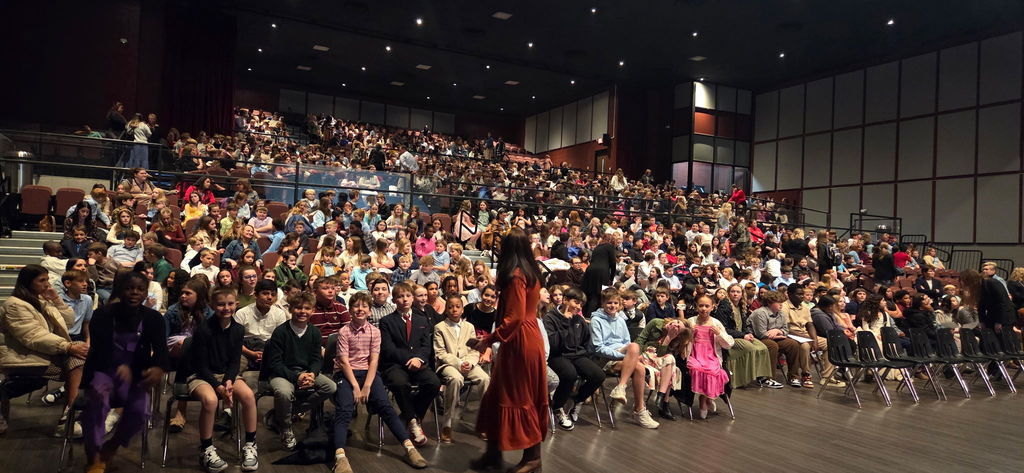 A wide view of a packed auditorium at the Citizens Center shows hundreds of fifth-grade students from across Lincoln County Schools seated in rows. Students in dressy casual attire fill the floor and tiered seating, chatting and waiting for the performance to begin.