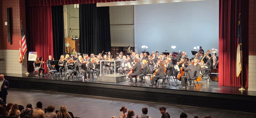 A full view of the North Carolina Symphony on stage at the Citizens Center shows musicians dressed in black performing under bright stage lights, with the American and North Carolina flags positioned on either side of the stage.