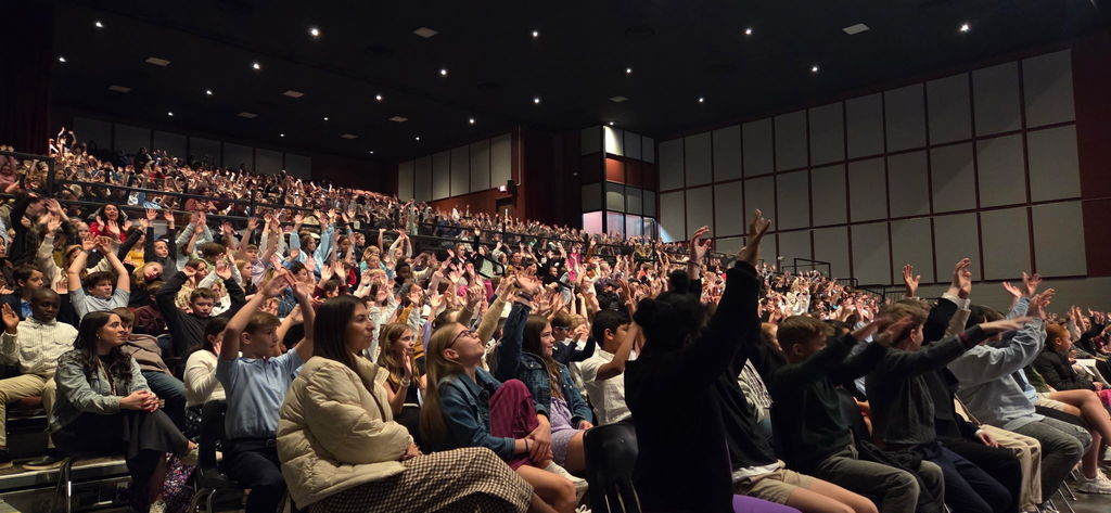 Hundreds of fifth-grade students seated in a large auditorium raise their hands high in the air as they participate in an interactive lesson about musical dynamics, learning the difference between forte and piano during the North Carolina Symphony performance.