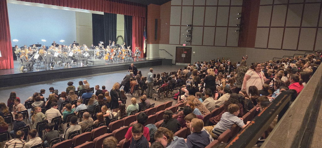 The North Carolina Symphony performs on stage at the Citizens Center, with musicians seated in sections playing string, brass, and percussion instruments. Fifth-grade students fill the audience, watching from the floor seats and balcony.