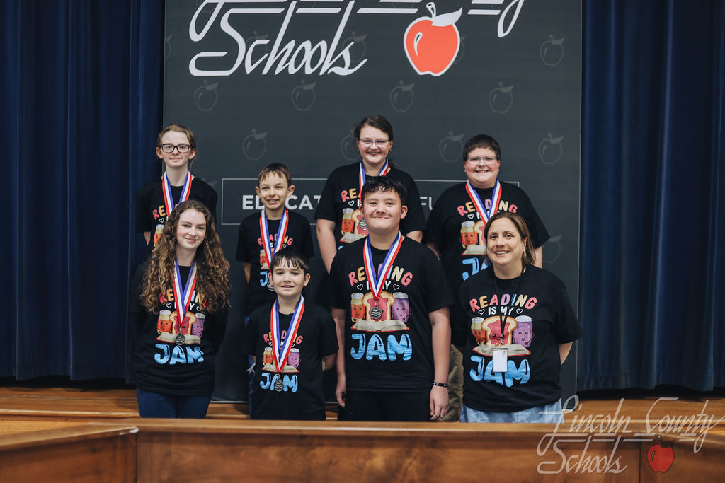 Middle school students wearing black “Reading Is My Jam” shirts pose with medals around their necks in front of a Lincoln County Schools backdrop. An adult coach stands at the side of the group.