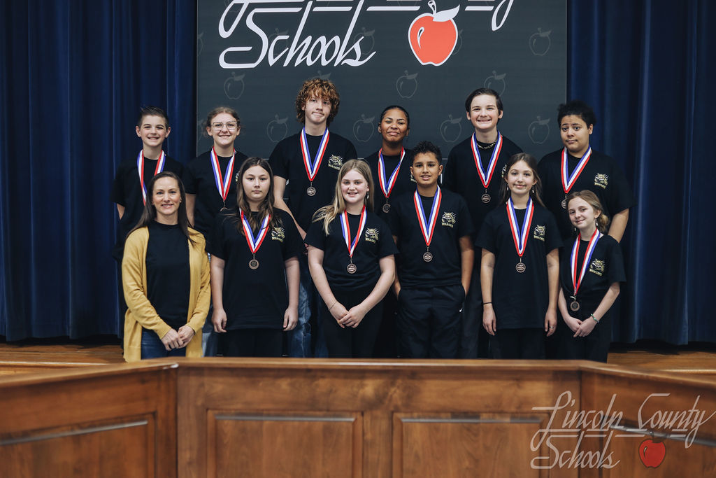 A team of middle school students dressed in black shirts with a school emblem stand together wearing medals. An adult coach stands beside them in front of a Lincoln County Schools backdrop.