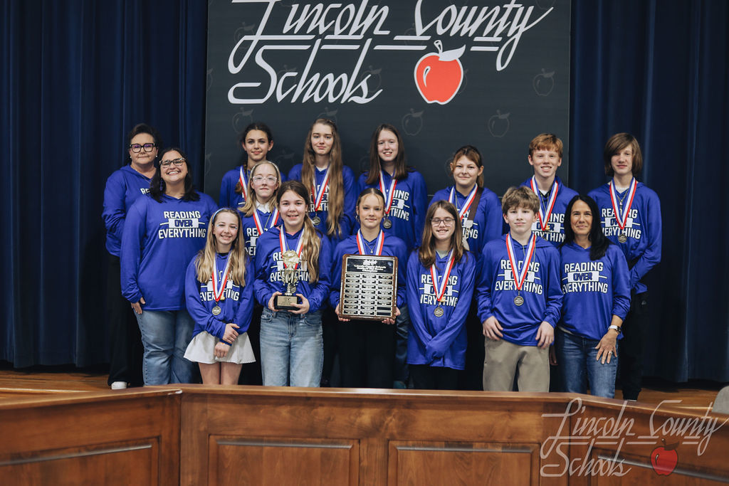 Group of middle school students wearing matching blue “Reading Over Everything” shirts stand in front of a Lincoln County Schools backdrop. Several students wear medals, and one student holds a large championship plaque while another holds a trophy. Two adult coaches stand on either side of the group.