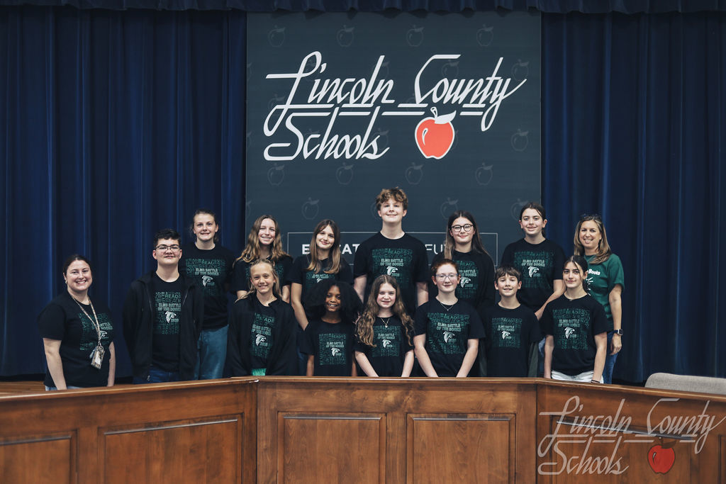 Another middle school team wearing dark green and black shirts pose together with medals in front of a Lincoln County Schools backdrop. Two adult coaches stand at each end of the group.