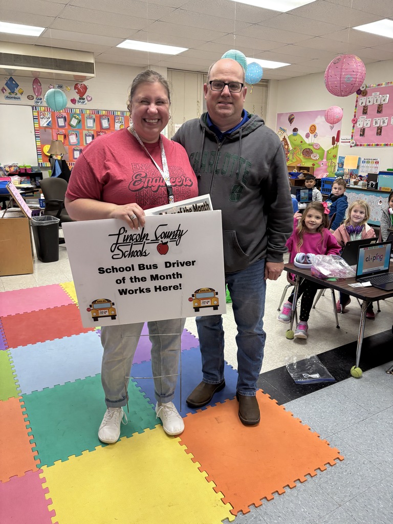 A smiling woman and man stand together in an elementary classroom. The woman holds a white sign that reads “Lincoln County Schools School Bus Driver of the Month Works Here!” Students sit at desks in the background. Colorful classroom decorations and hanging paper lanterns are visible.