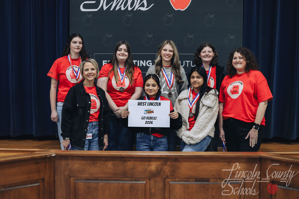 West Lincoln High School students and coaches wearing red shirts and medals pose on stage. A student in front holds a sign reading “West Lincoln – Go Rebels! – 2026.”