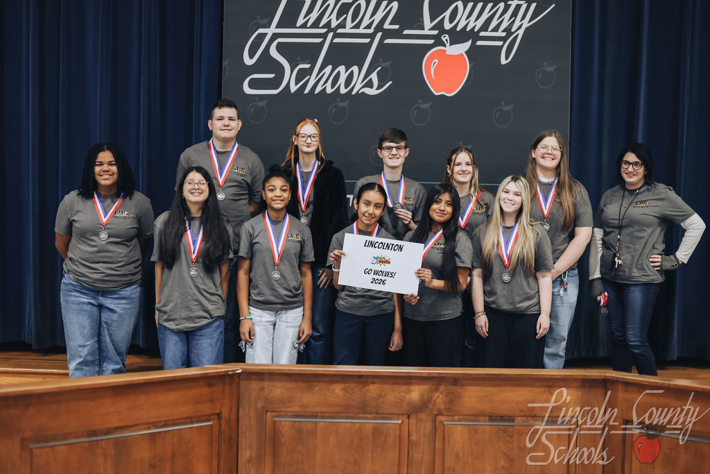 Lincolnton High School students wearing gray shirts and medals stand together on stage. A student in the center holds a sign reading “Lincolnton – Go Wolves! – 2026.”