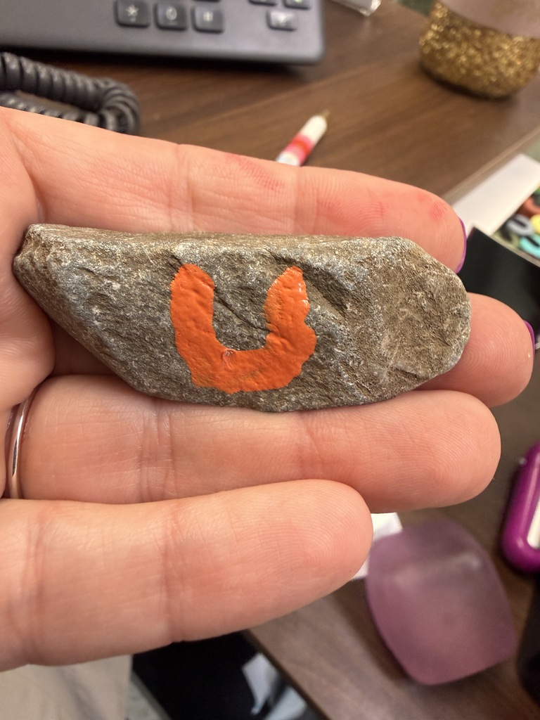 Close-up of a hand holding a small gray rock with an orange painted letter shaped like a “U.” The rock rests across the person’s fingers, with a desk surface, keyboard, and office items blurred in the background.
