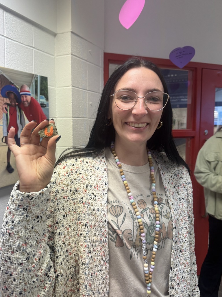 A woman with long dark hair and glasses smiles while holding up a small rock painted with an orange letter. She wears a patterned cardigan, a graphic T-shirt, and a beaded lanyard necklace. The background shows a school hallway with red-framed windows and heart-shaped decorations on the wall.