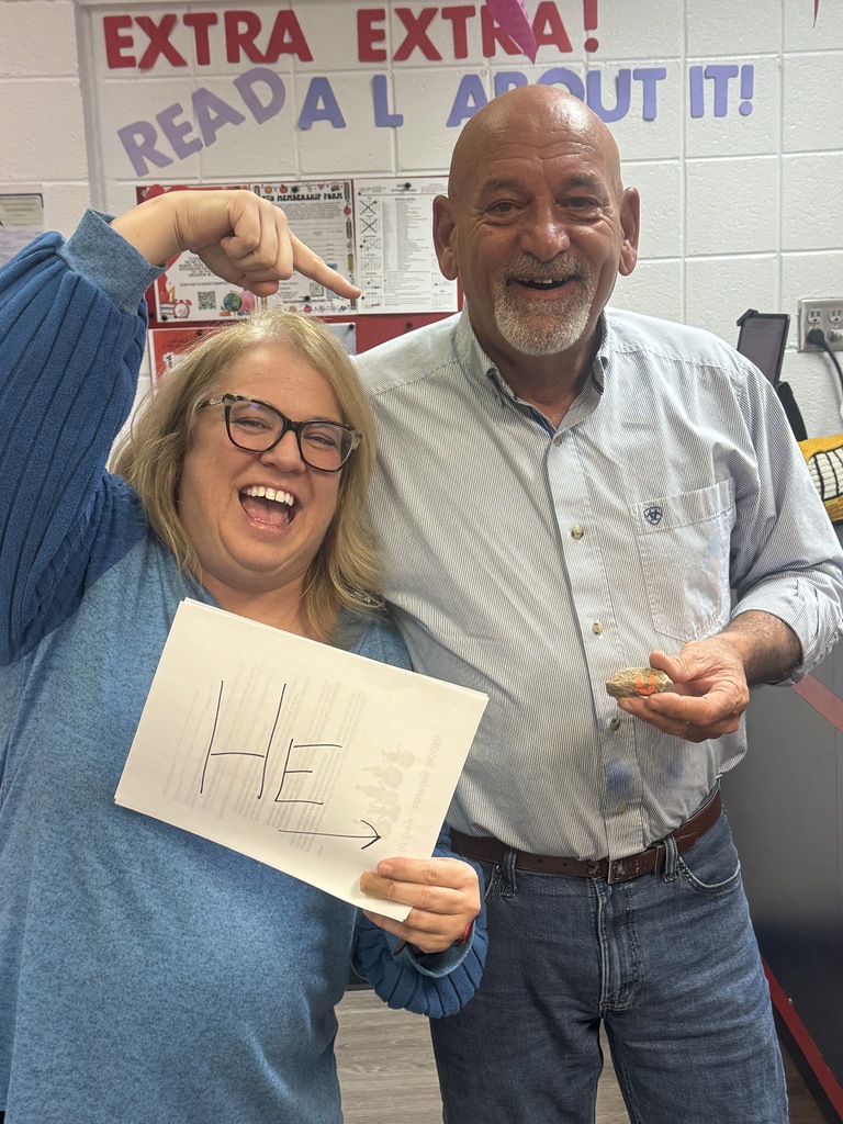 A smiling woman in a blue sweater holds a sheet of paper with the letters “HE” and an arrow written in black marker while pointing playfully toward a bald man standing beside her. The man smiles and holds a small rock with an orange painted letter on it. A classroom wall behind them displays a sign that reads “EXTRA EXTRA! READ ALL ABOUT IT!” along with posted papers and decorations.