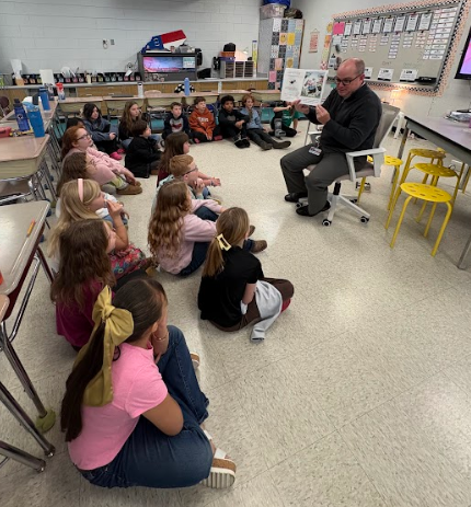 A guest reader sits in a chair at the front of a classroom, reading a book to a group of students gathered on the floor in front of him
