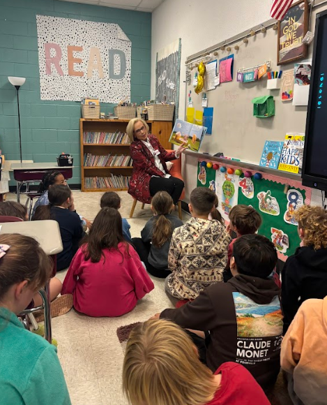 A guest reader in a red patterned jacket sits on a chair in a classroom, reading a picture book to a group of elementary students who are sitting on the floor around her.