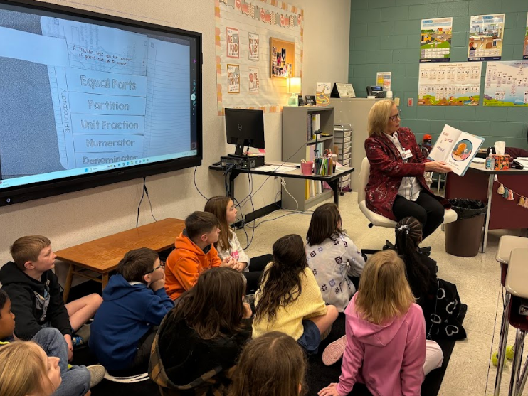 A woman in a red jacket reads the book Peanut Butter & Cupcake! to a group of students seated on a rug