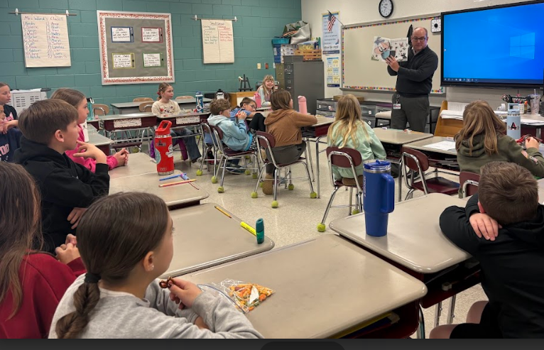 A male guest reader stands at the front of a classroom, holding up a picture book for students who are seated at their desks. A large blue digital screen is mounted on the wall behind him.