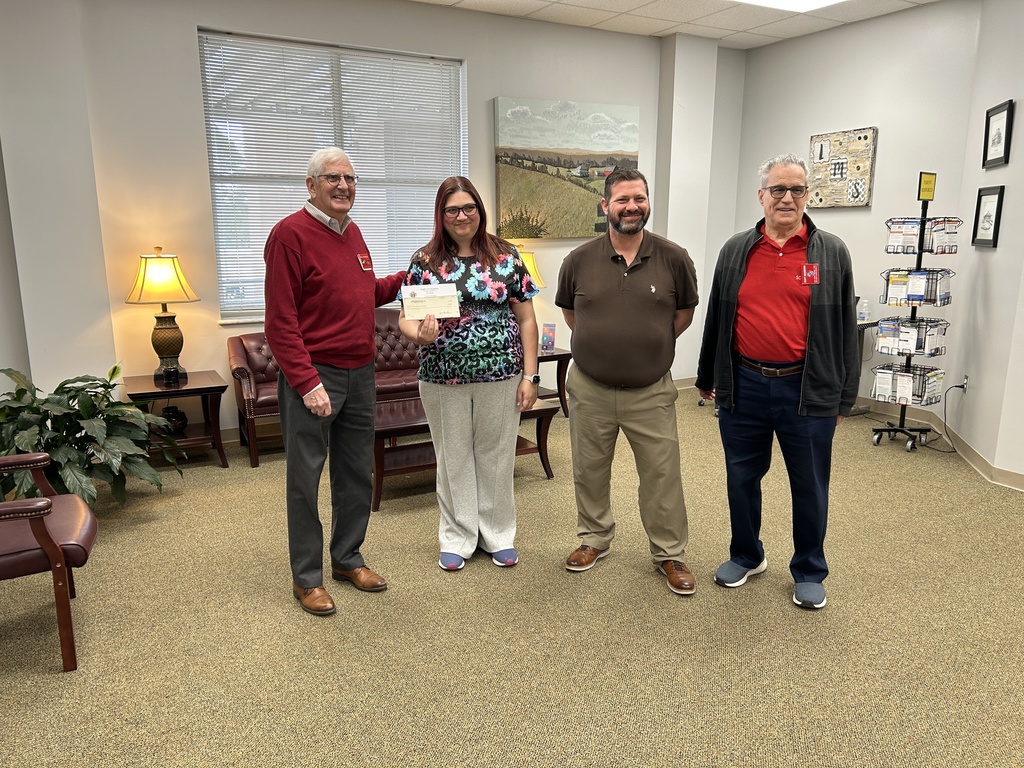 Four adults stand in a professional office or lobby setting with tan carpeting and light-colored walls. On the far left, an older man in a red sweater and grey slacks stands next to a woman wearing a floral and leopard print scrub top and light grey pants. The woman is holding a white envelope or small document. To her right stands a man in a brown polo shirt and khaki pants. On the far right, a man wears a red polo shirt under a dark zip-up jacket with navy blue trousers. In the background, there is a brown leather sofa, a wooden coffee table, a large landscape painting, and a wire rack containing brochures.