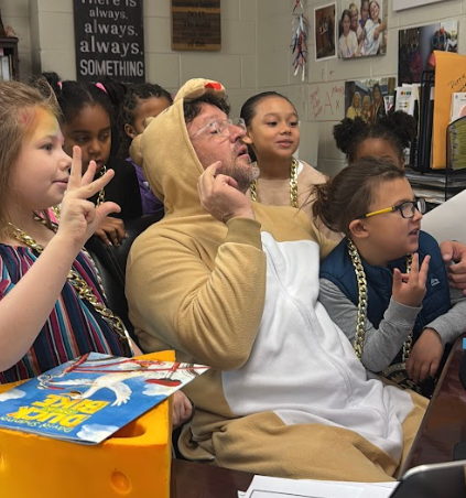 A group of elementary students crowd around an adult wearing a brown bear costume and oversized gold chain necklace. The adult sits at a desk with a laptop open while students smile and hold up hand signs. A children’s book rests on a yellow box in the foreground, and classroom posters and photos are visible on the wall behind them.