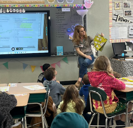 A teacher sits at the front of a classroom holding up a colorful picture book while students sit at desks facing her. An interactive board displays a document with highlighted text, and classroom decorations and learning materials line the walls.