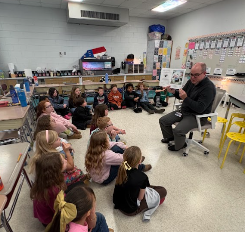 An adult male sits on a chair at the front of a classroom, holding open a picture book and reading to a group of elementary students seated cross-legged on the floor. Classroom shelves, student desks, and bulletin boards surround the group as students listen closely.