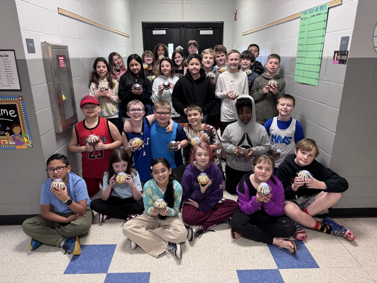 A large group of approximately twenty-five fifth grade students gathered in a school hallway. The students are arranged in three rows: some are sitting on the blue and white tiled floor, while others stand behind them. Nearly every student is smiling and holding a small, individually wrapped bundt cake. They are dressed in casual clothing, including t-shirts, hoodies, and athletic jerseys. The background features cinder block walls, a doorway at the end of the hall, and classroom signs labeled C-8 and C-7.