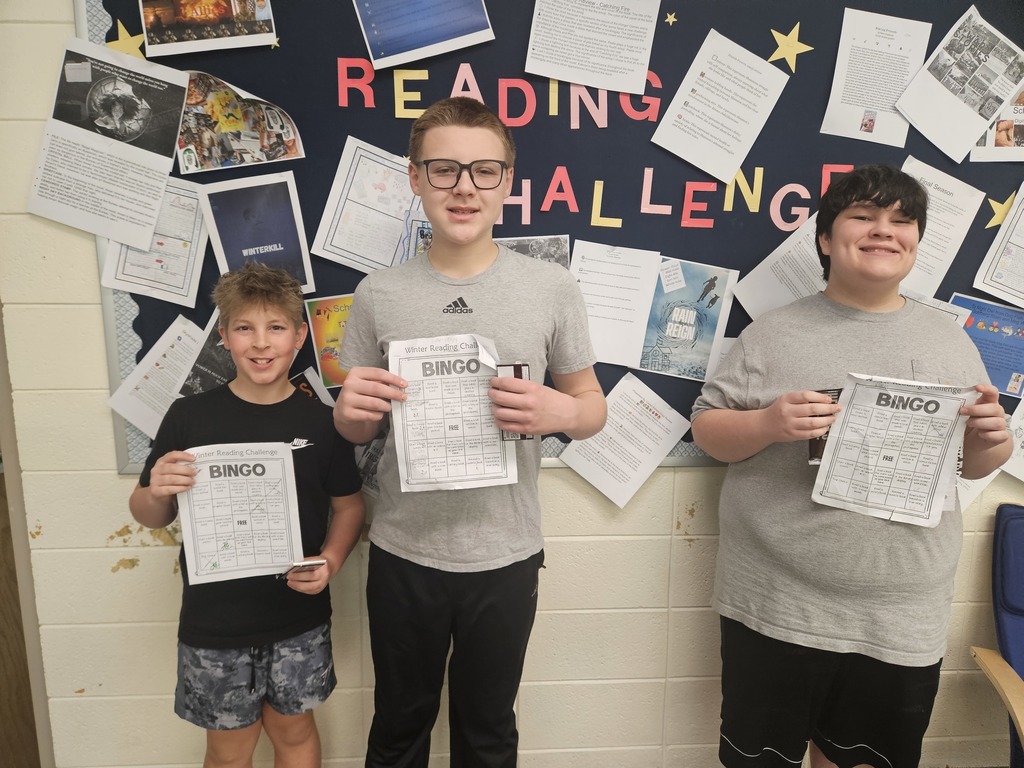 Three boys pose together in front of the same "READING CHALLENGE" bulletin board, displaying their completed "Winter Reading Challenge BINGO" sheets and chocolate bar prizes. The boy on the left wears a black Nike t-shirt and patterned shorts, the center boy wears a grey Adidas t-shirt and glasses, and the boy on the right wears a plain grey t-shirt and black shorts. The wall behind them features a mix of printed book reports and student projects pinned to a blue background with a white scalloped border.