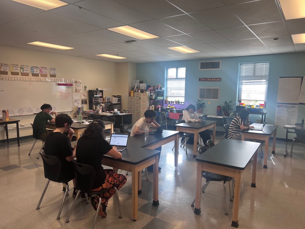 A classroom setting where six students are working at black-topped lab tables. The room has light yellow and blue walls, with "Mrs. Beckham" displayed on a sign above an air vent. Most students are focused on laptops, while one student in the foreground wears orange patterned pants and another in the back looks toward the camera. Sunlight filters through two windows with blinds, and the walls are decorated with educational posters and a whiteboard.