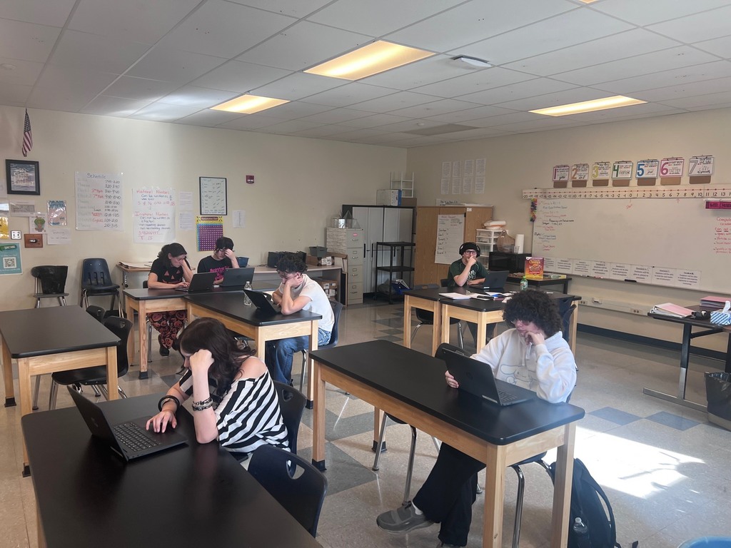 An indoor shot of a classroom with six students sitting at black-topped lab tables, all focused on their laptops. The room features white walls decorated with various educational posters, a daily schedule, and a long whiteboard with number lines and notes across the back wall. The students are spread throughout the room, some wearing headphones, while the overall atmosphere appears quiet and studious.