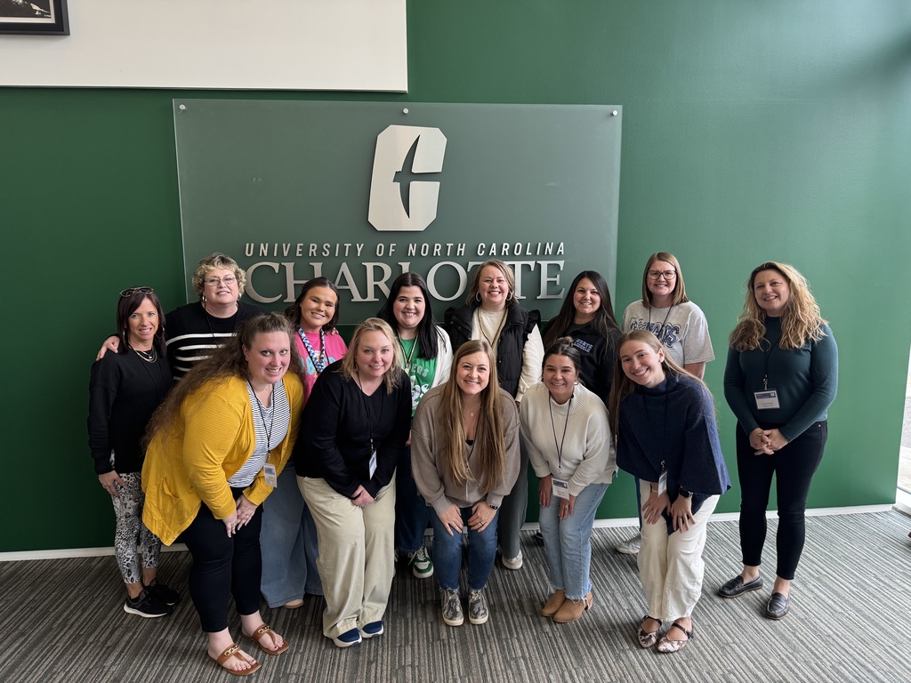 A group of Lincoln County Schools teachers pose together in front of a green wall sign that reads “University of North Carolina Charlotte.” The group stands and kneels in two rows, smiling for the photo during their visit to the SWEA Literacy Conference at UNC Charlotte.