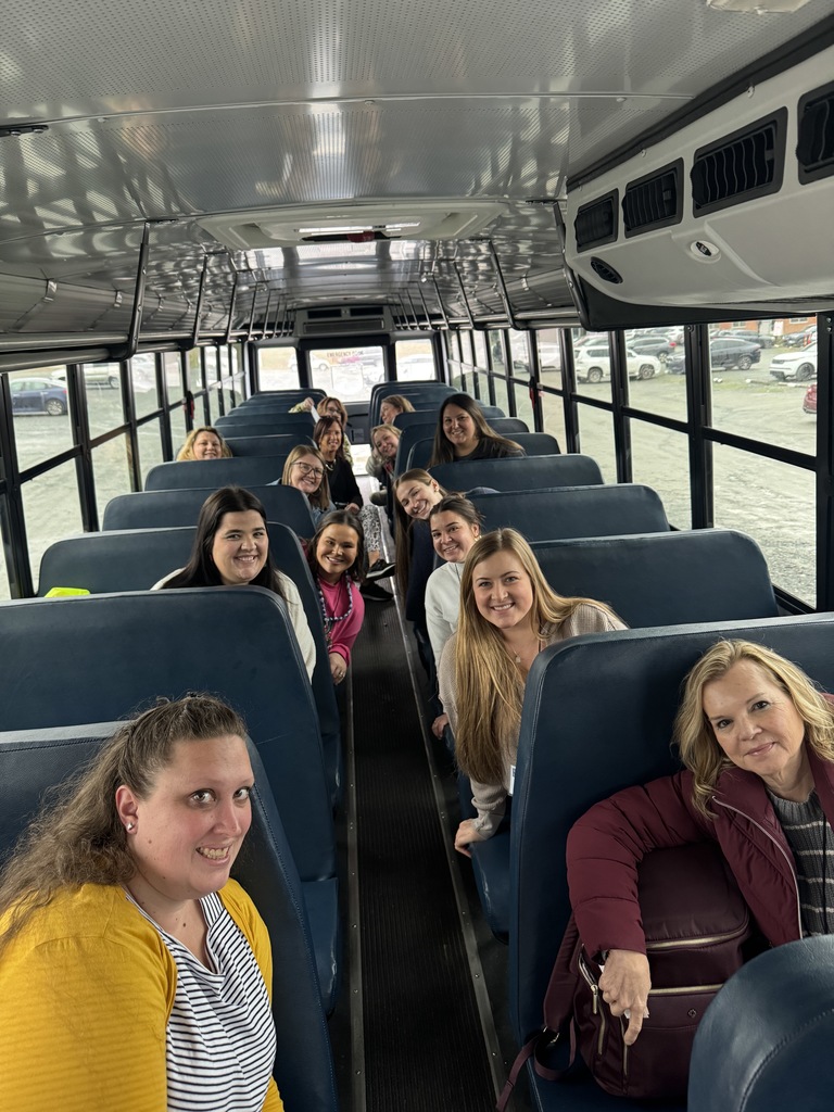 Lincoln County Schools teachers sit on a school bus smiling toward the camera while traveling together to attend the SWEA Literacy Conference at UNC Charlotte. The group fills several rows of seats inside the bus.