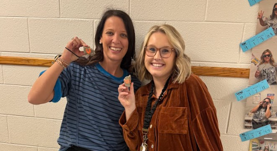 Two women stand against a hallway wall, smiling and holding up small rocks painted with orange “U” letters. One wears a striped blue shirt and the other wears glasses and a brown jacket with a lanyard. Student photos and decorations are displayed on the wall behind them.