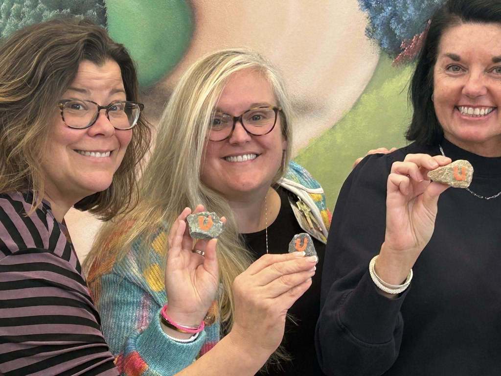 Three women stand close together smiling while holding small gray rocks painted with bright orange “U” letters. They pose in front of a colorful mural background, each holding their rock up near their faces so the painted letter is clearly visible.