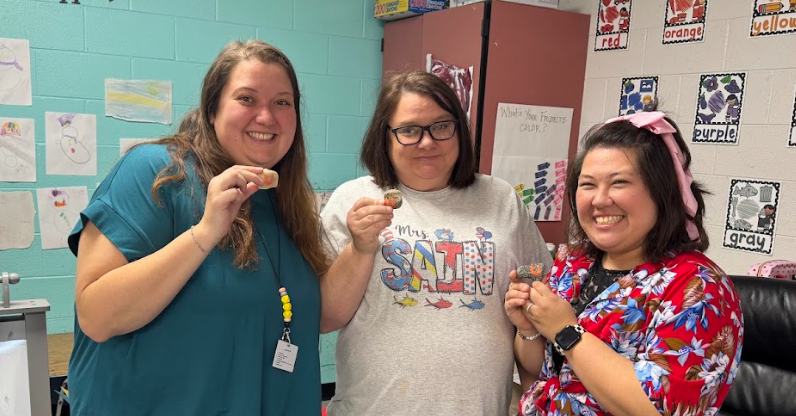 Three women stand together inside a classroom, smiling while holding small rocks painted with orange “U” letters. Behind them are classroom posters showing color words like “purple,” “red,” and “yellow,” along with student drawings displayed on the wall.