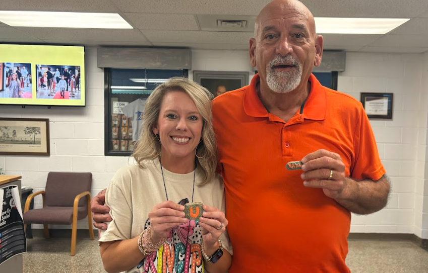 A man wearing an orange polo shirt stands beside a smiling woman in a school office or lobby area. Both hold small rocks painted with orange “U” letters. The man has his arm around the woman’s shoulder while framed photos and a television screen are visible on the wall behind them.