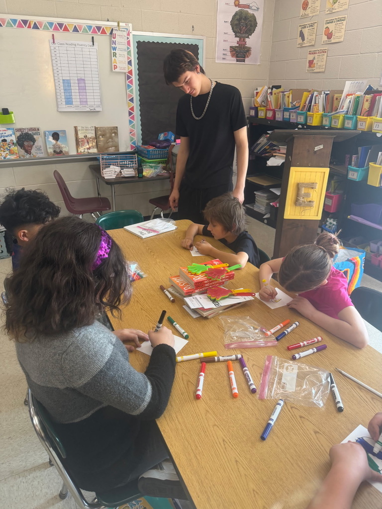 Gemini said A young man in a black t-shirt and silver chain stands over a wooden classroom table, observing four younger students as they draw with markers. The students are focused on their small illustrations, with markers and plastic bags scattered across the table. In the background, there is a whiteboard with a reading fluency chart, bookshelves filled with books, and a wooden podium.