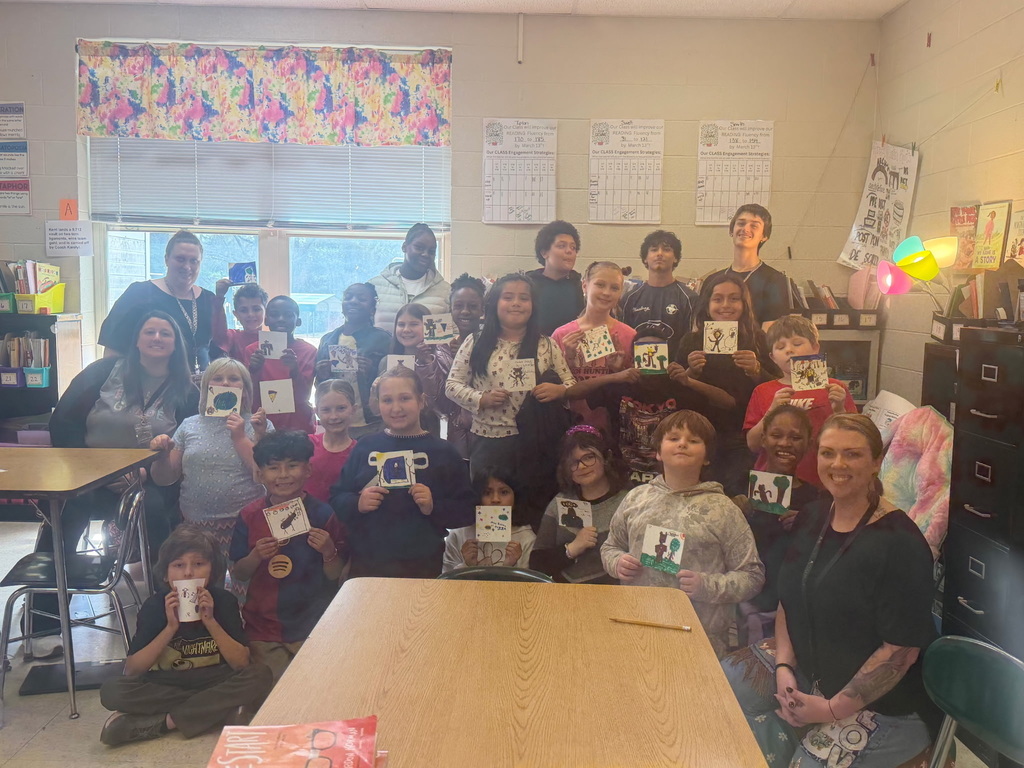 A large group of about twenty students and three teachers posing for a photo in a classroom. Most of the students are standing or kneeling and proudly holding up small, colorful square drawings they have created. The room is filled with classroom materials, including posters on the walls, books on shelves, and a large wooden table in the foreground. Everyone is smiling or looking toward the camera, capturing a moment of collective pride in their creative work.