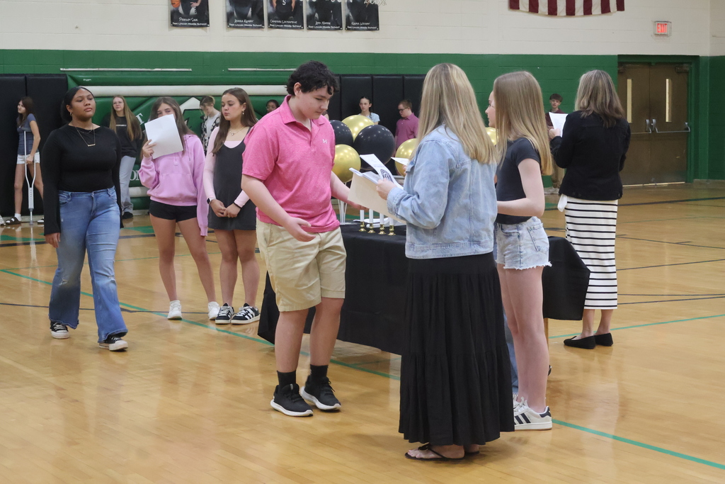 In a high school gymnasium, an adult woman stands at a table draped in black, leading a ceremony while reading from a document. She is joined by several teenagers, some of whom are holding scripts or receiving papers, as others wait in a line along the green and white walls in the background. The scene is decorated for a formal school event with black and gold balloon clusters and small white candles on the table, while a "Falcon Pride" banner and a falcon mascot logo emphasize the school's identity.