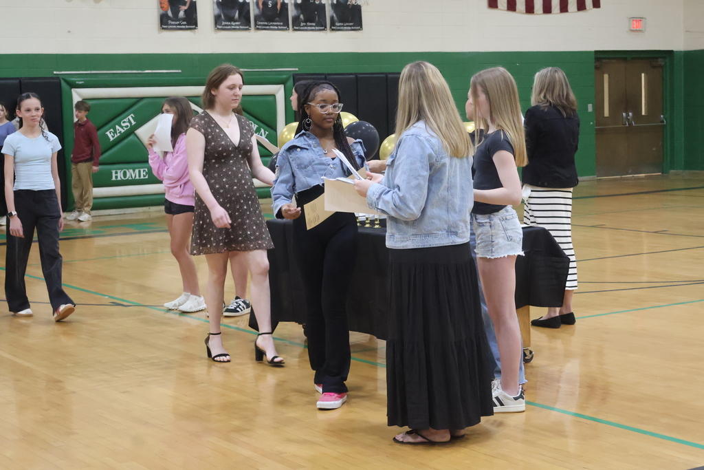 In a high school gymnasium, students participate in a formal induction ceremony at a table draped in black and decorated with gold and black balloons and white candles. Adult facilitators and student leaders stand at the table to read from documents and distribute certificates or programs to a line of teenagers dressed in a mix of casual and semi-formal attire. The background features green and white walls adorned with "Falcon Pride" banners, a falcon mascot logo, and athletic posters, capturing the school spirit of the event.