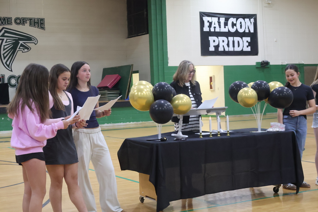 In a high school gymnasium, an adult woman stands behind a table draped in black, leading an induction or ceremony while reading from a paper. To her left, three teenage girls stand together holding scripts or programs, while another girl stands to the right of the table. The setting is decorated with black and gold balloon clusters and small white candles on the table, with a "Falcon Pride" banner and a falcon mascot logo visible on the green and white walls in the background.