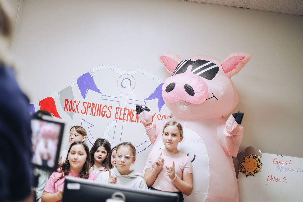 A group of elementary students stand in front of a Rock Springs Elementary School backdrop while posing with a large pink inflatable pig wearing sunglasses. A tablet on a tripod records the group as they hold their hands up in playful poses.