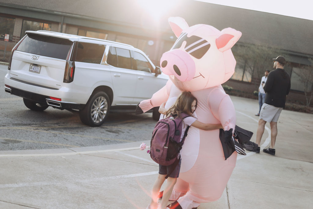 A student wearing a backpack hugs a large pink inflatable pig mascot outside a school building during morning arrival, while cars and adults stand nearby in the pickup area.
