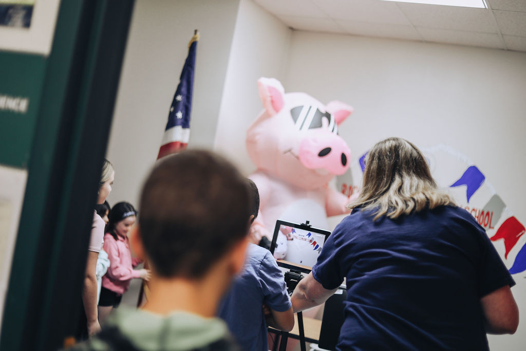 Students gather in a small room while an adult films a large inflatable pig mascot standing in front of a Rock Springs Elementary School banner. The scene is viewed from behind several students watching the recording.
