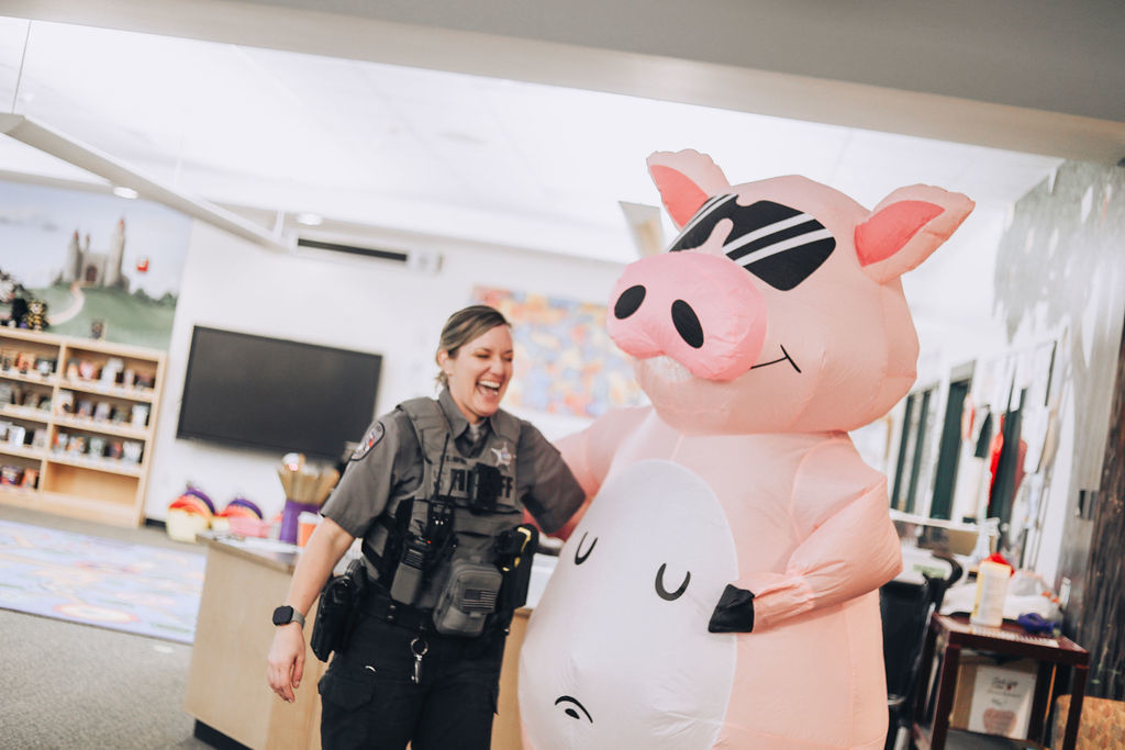 A school resource officer laughs while walking beside a large pink inflatable pig mascot inside a school media center. Bookshelves and classroom displays appear in the background.