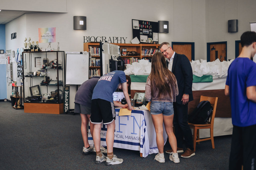 Students stand at a financial management booth while one student writes on a form and an adult representative speaks with them about career opportunities in the library.
