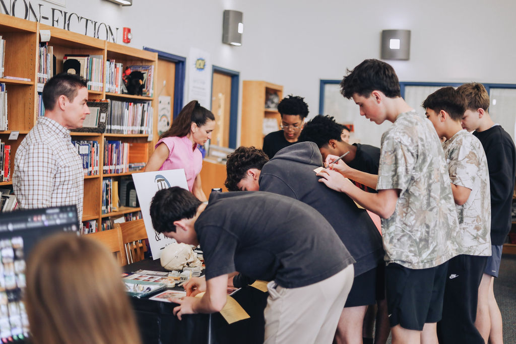 A group of high school students lean over a table display, filling out forms and examining items while talking with a presenter during a career exploration event in the school library.