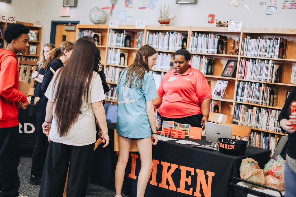 Students stand around a Timken display table set up in a school library, speaking with a representative and looking at materials about careers while bookshelves line the background.