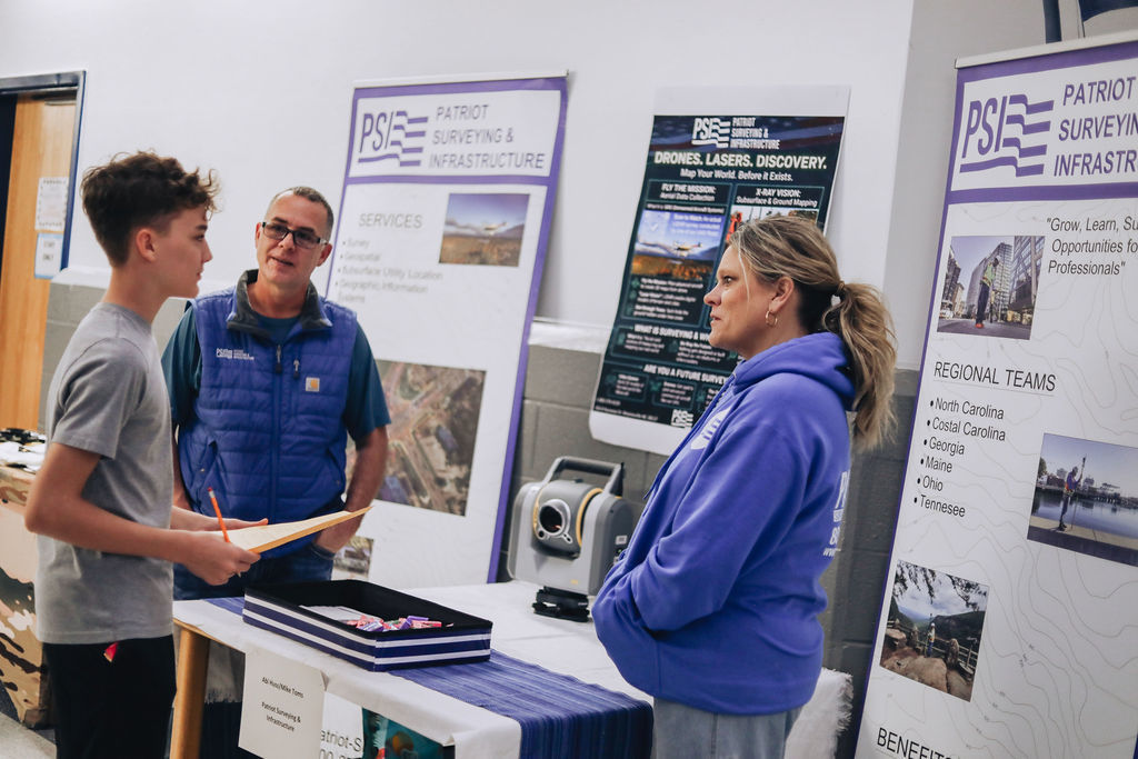 A student holding a clipboard talks with two representatives at a Patriot Surveying and Infrastructure display booth, surrounded by informational posters and equipment used in surveying careers.