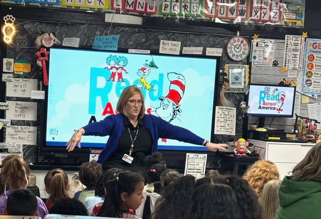 A teacher stands in front of a classroom full of students with a screen displaying “Read Across America” and Dr. Seuss characters while she gestures with her arms during a reading activity.