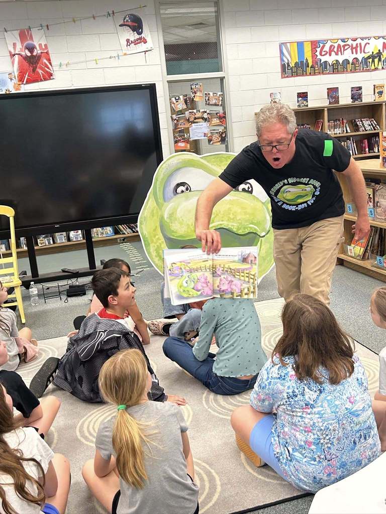 A guest reader animatedly shows a picture book to a group of elementary students sitting together on a rug in the media center, with a large green frog decoration and bookshelves visible in the background.