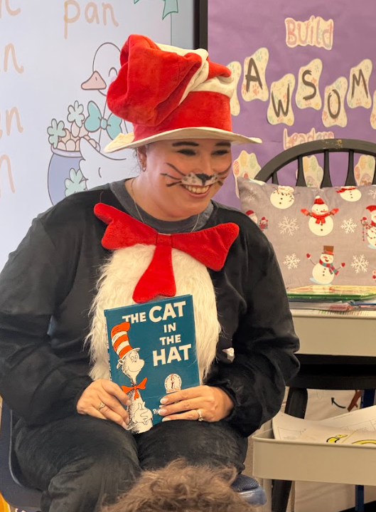 A teacher dressed as the Cat in the Hat smiles while holding a copy of The Cat in the Hat book as she reads to students during a themed classroom reading activity.