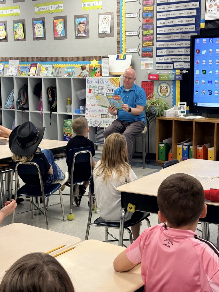 An adult guest reader sits at the front of an elementary classroom reading a picture book to students seated at their desks. Colorful classroom charts, student artwork, and a word wall are displayed behind him.